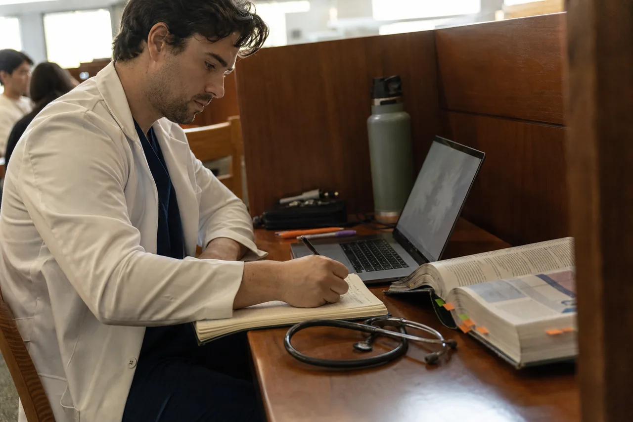 Physician assistant trainee comparing two study subscriptions on a laptop beside a pharmacology textbook in a campus library